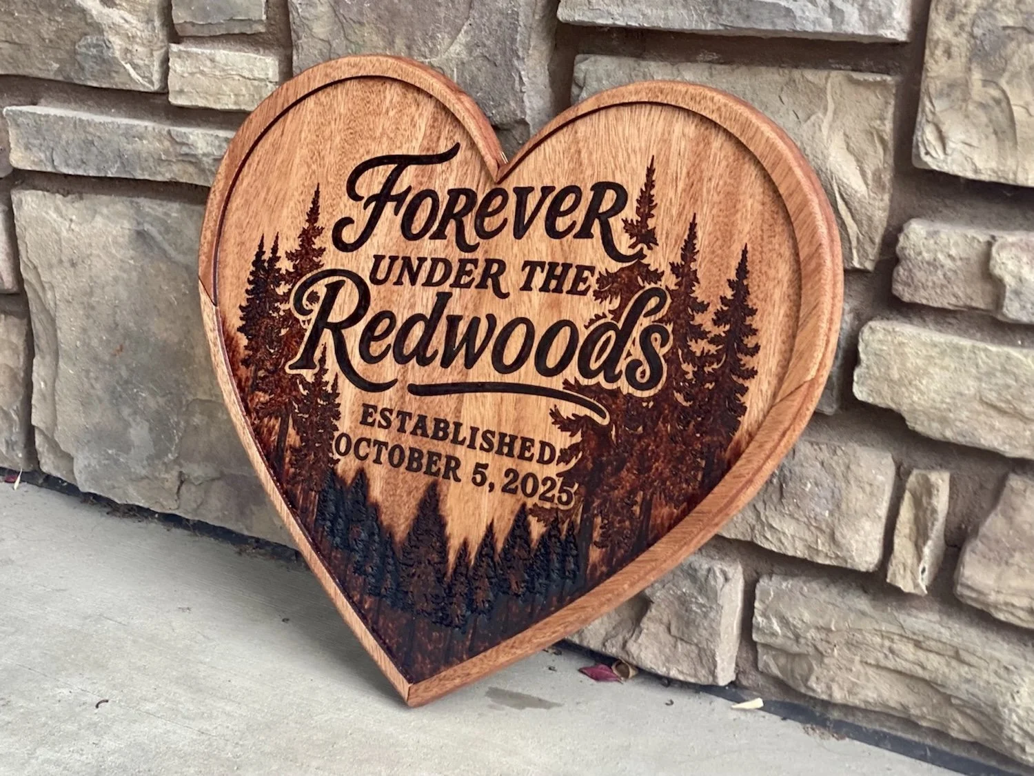 A wooden heart-shaped plaque with engraved text that reads 'FOREVER UNDER THE REDWOODS' and 'ESTABLISHED OCTOBER 5, 2025,' featuring engraved images of redwood trees, placed against a stone wall.
