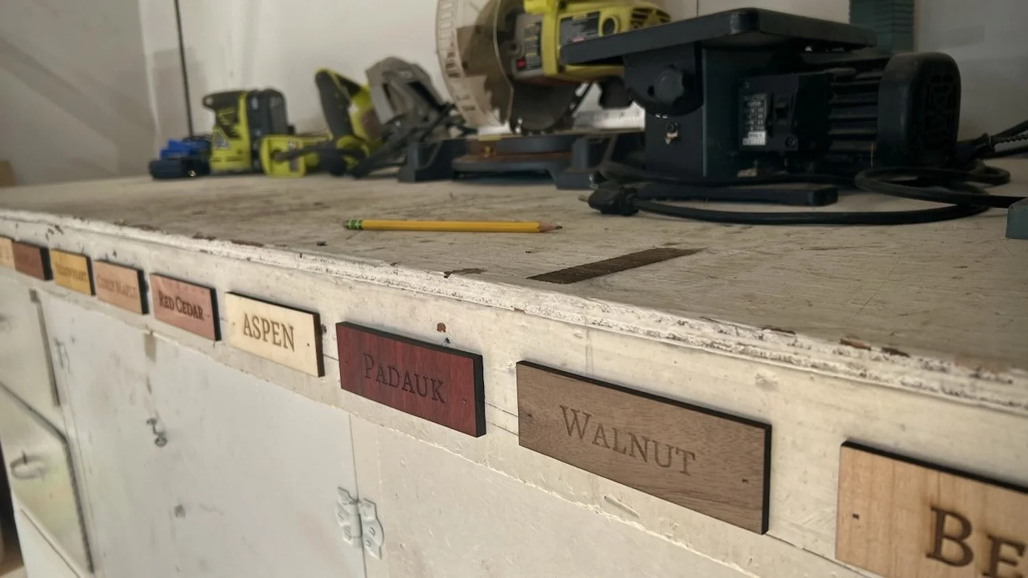 A worn workshop desk with various power tools and a yellow pencil on top. Small labeled wood strips are attached to the front edge of the desk, displaying names like Aspen, Padauk, Walnut, and Cedar.