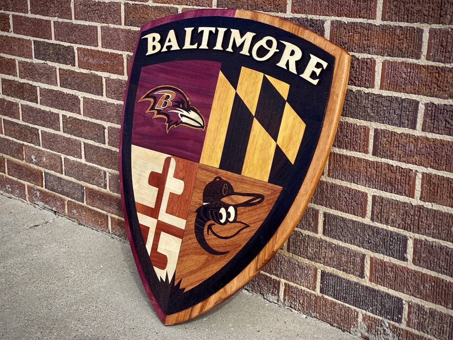 A decorative wooden shield featuring the Baltimore Ravens logo, Maryland state flag, a cartoon duck wearing a baseball cap, and a Christian cross, leaning against a brick wall.