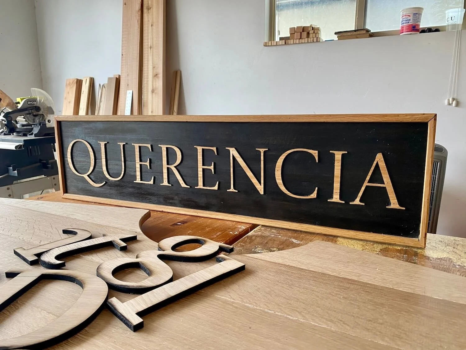 A wooden sign with the word 'QUERENCIA' carved into it, placed on a workbench in a woodworking shop.