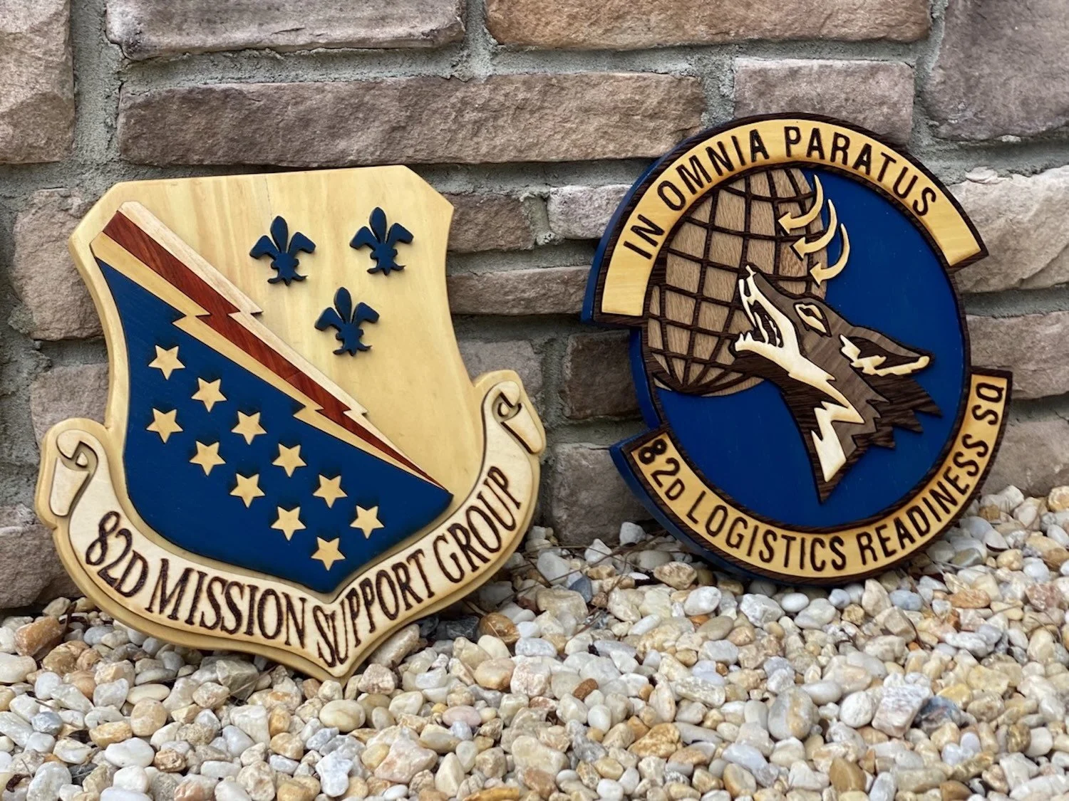 Two military patches on a brick wall background. The left patch is shaped like a shield with blue and gold colors, stars, and a lightning bolt The right patch features a wolf howling at a globe.