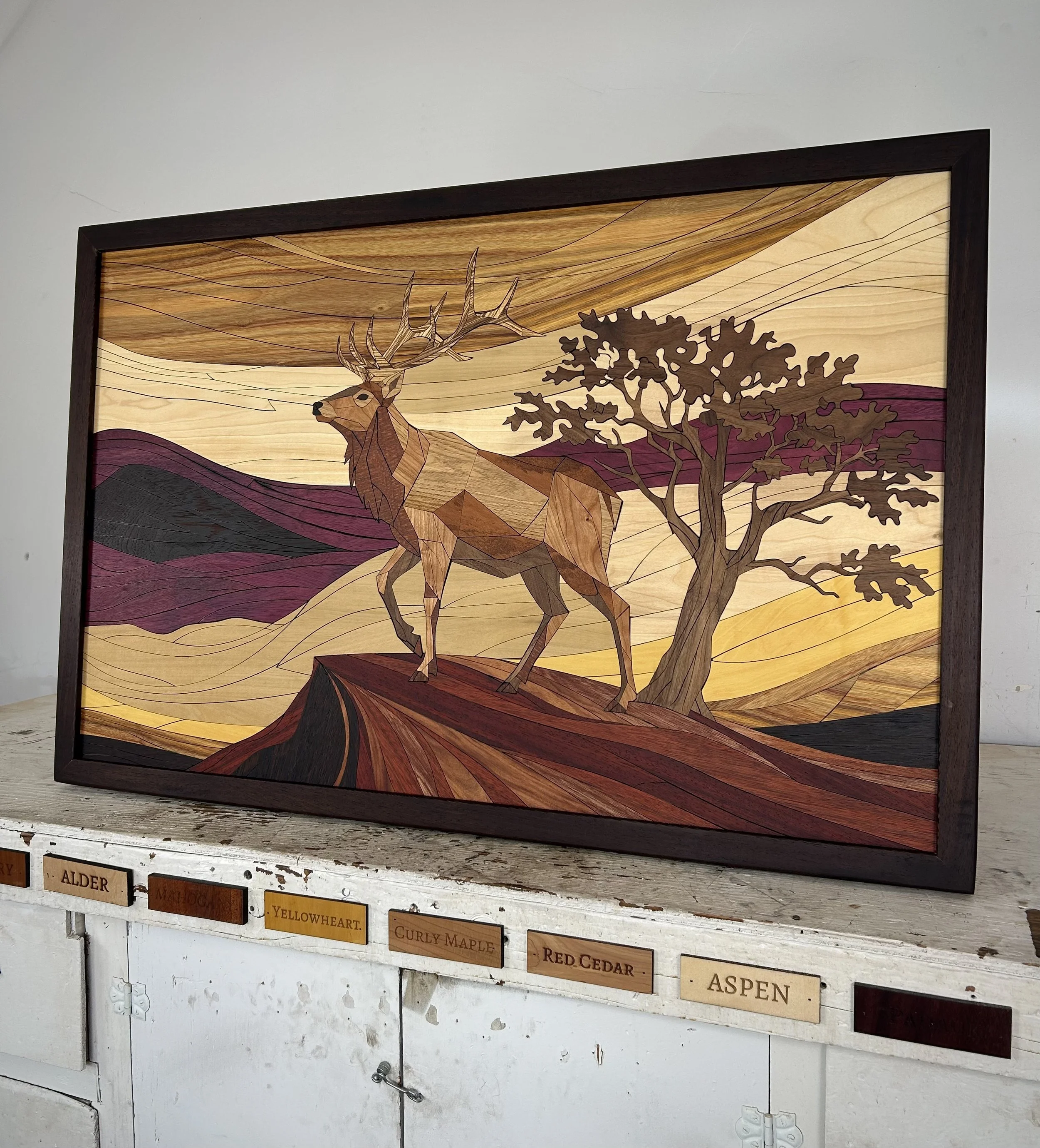 Wooden artwork featuring a geometric deer standing next to a tree, with layered mountains and a sky in the background. The scene is framed with a dark wooden border.