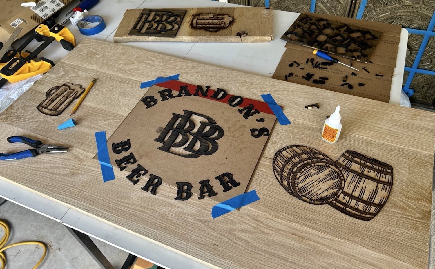 A woodworking project in progress with a sign for 'BRANDON'S BEER BAR' featuring a logo with stylized 'BB' initials, various wood and metal tools, a bottle of glue, and several sketches and cutouts of barrels on a worktable.