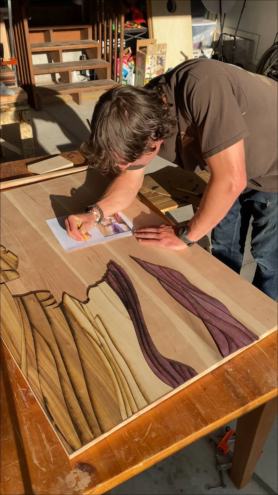 A man is leaning over a wooden table, working on a wood art piece that features various pieces of wood arranged in a flowing pattern, with a comic book page placed on the table.