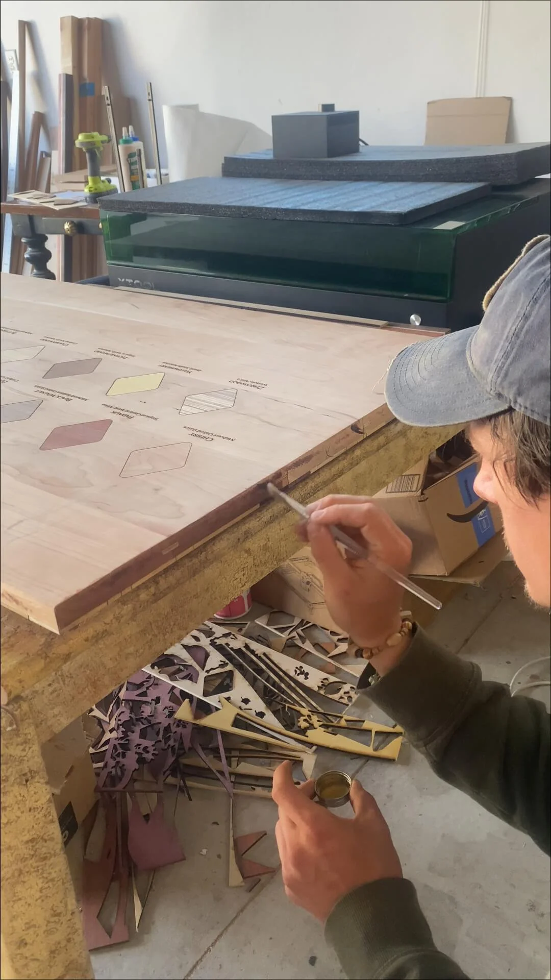 Person working on a woodworking project, sitting at a workbench with various colored wood samples, tools, and cutout wood pieces scattered around.