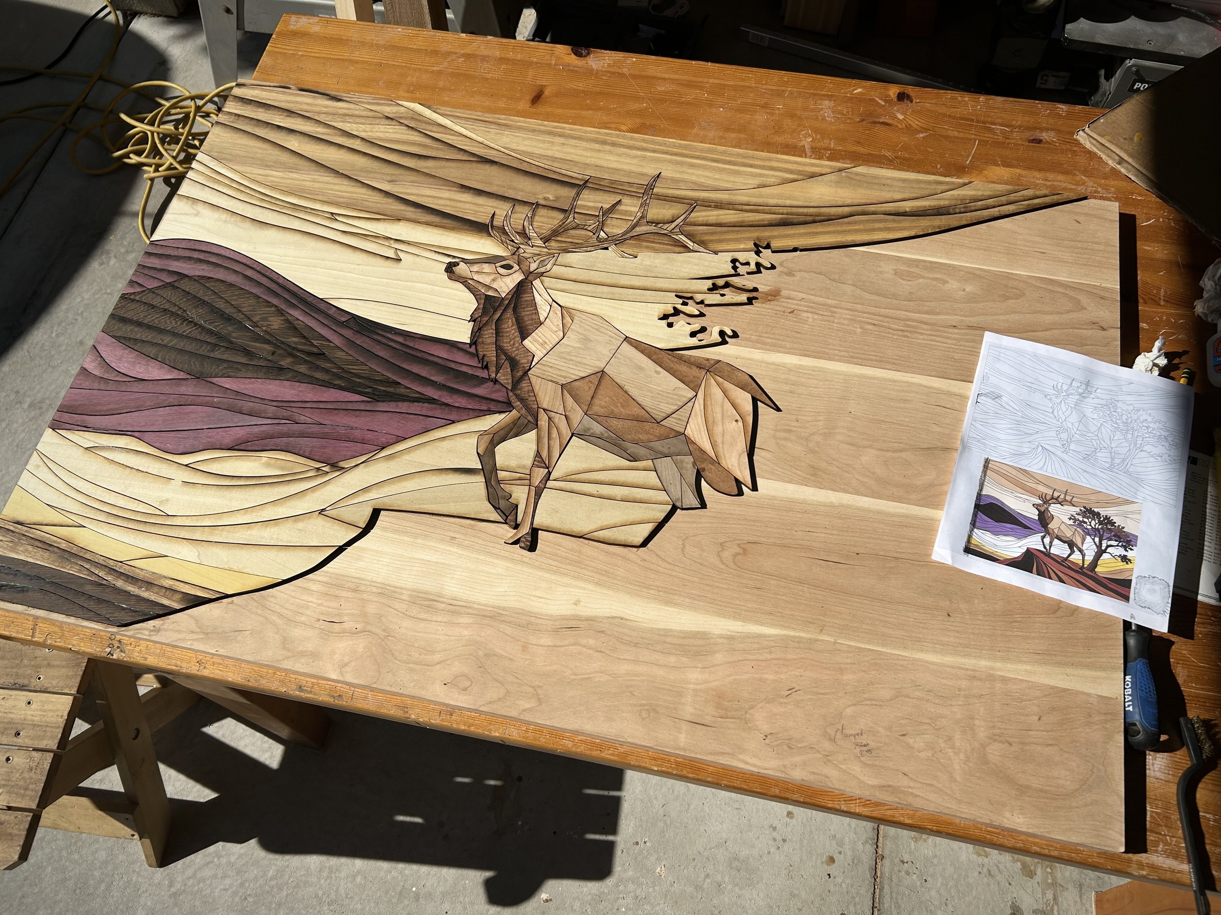 A wooden artwork depicting a deer with large antlers, surrounded by stylized flowing lines that resemble a landscape. There is a printed version of the same artwork and a sketch on the right side of the table.