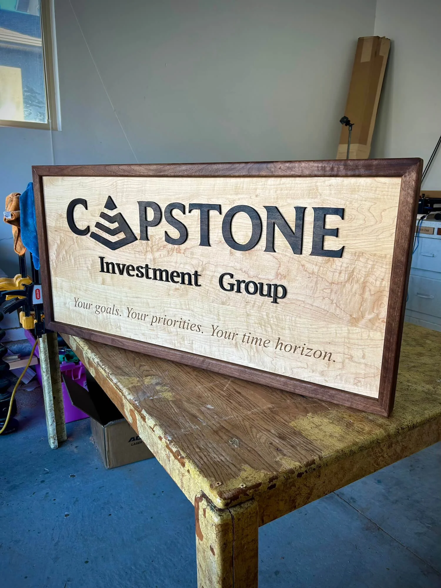 Wooden sign for Capstone Investment Group with a logo, tagline, and motivational message on a workbench in a workshop.