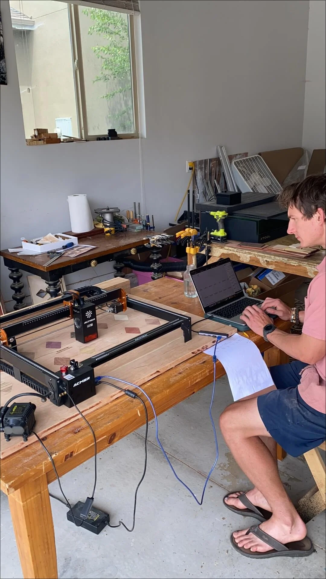 A man working with a laptop and 3D printer at a wooden workbench in a workshop.
