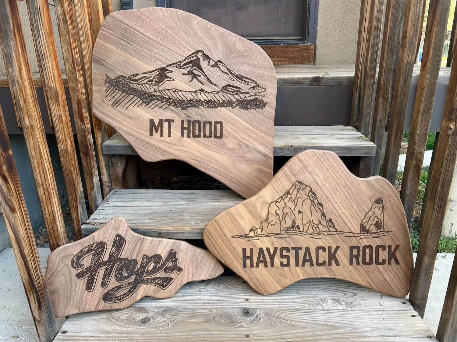 Three wooden signs with mountain and rock designs. One says "MT HOOD," another says "HAYSTACK ROCK," and the third says "Hops." They are leaning against a rustic wooden bench and railing.