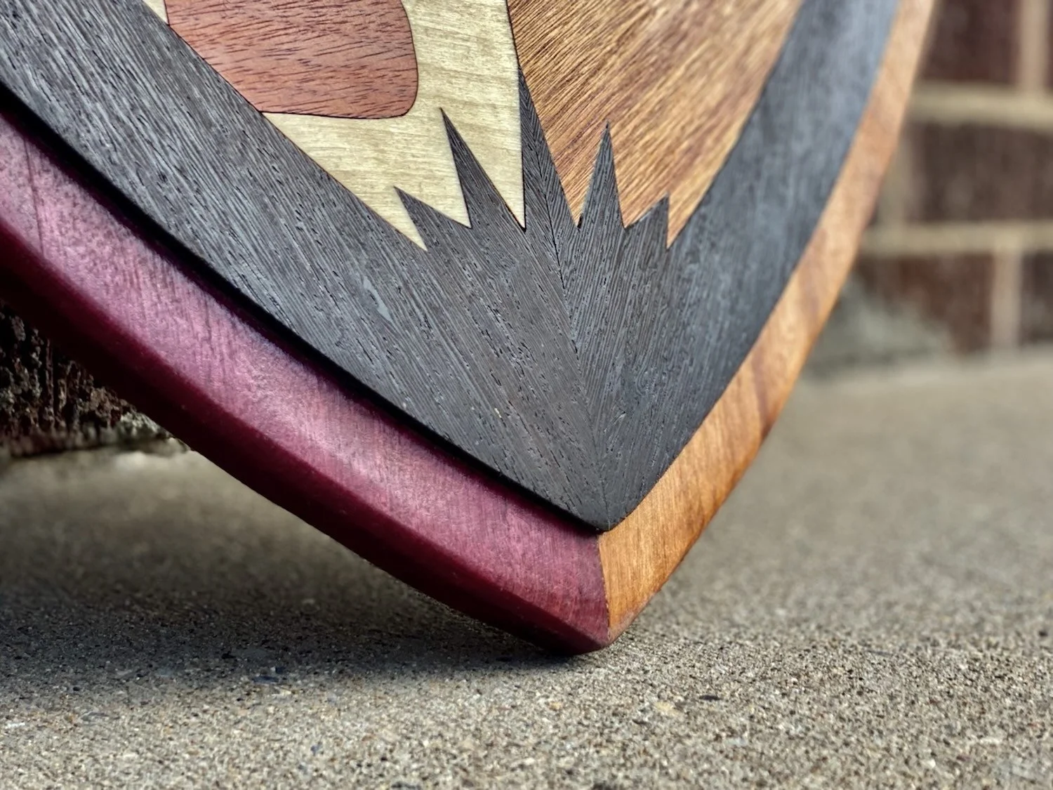 Close-up of a corner of a wooden artwork with varied wood tones, resting on concrete ground.