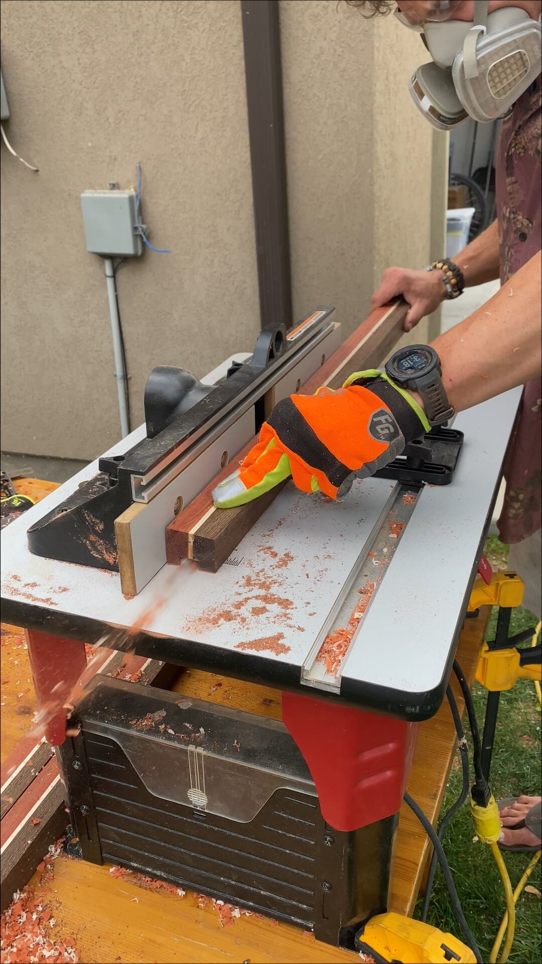 A person wearing gloves and a wristwatch is using a miter saw to cut a piece of wood on a table saw in an outdoor workshop.