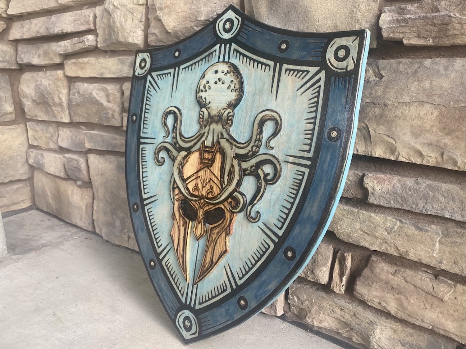 A decorative wooden shield mounted against a stone wall. The shield features a design of an octopus intertwined with a helmet. The shield has a blue and beige color scheme with black outlines.