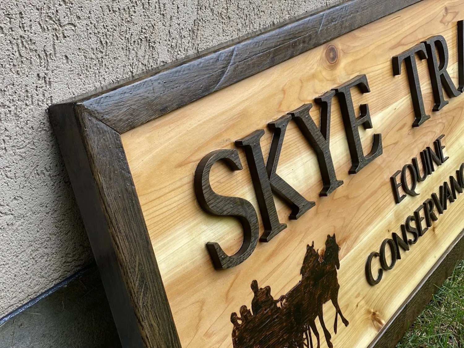 Close-up of a wooden sign with raised black letters that reads 'Skye Trace".  The sign has a natural wood finish with darker wood framing and an woodburning of a horse-drawn carriage with horses.