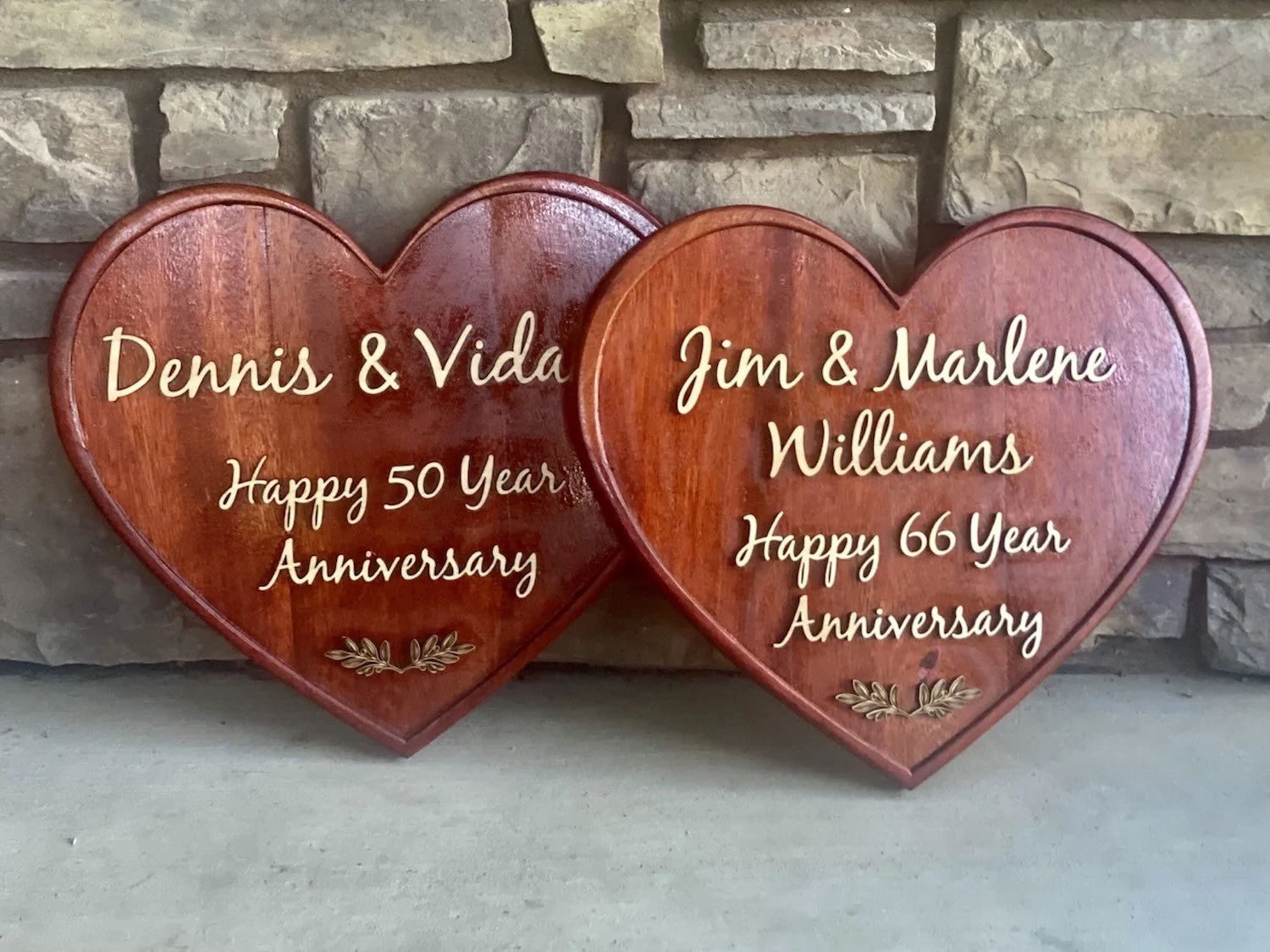 Two wooden hearts with engraved text celebrating wedding anniversaries, placed on a concrete surface in front of a stone wall. The left heart reads, "Dennis & Vida, Happy 50 Year Anniversary," and the right heart reads, "Jim & Marlene Williams, Happy 66 Year Anniversary."