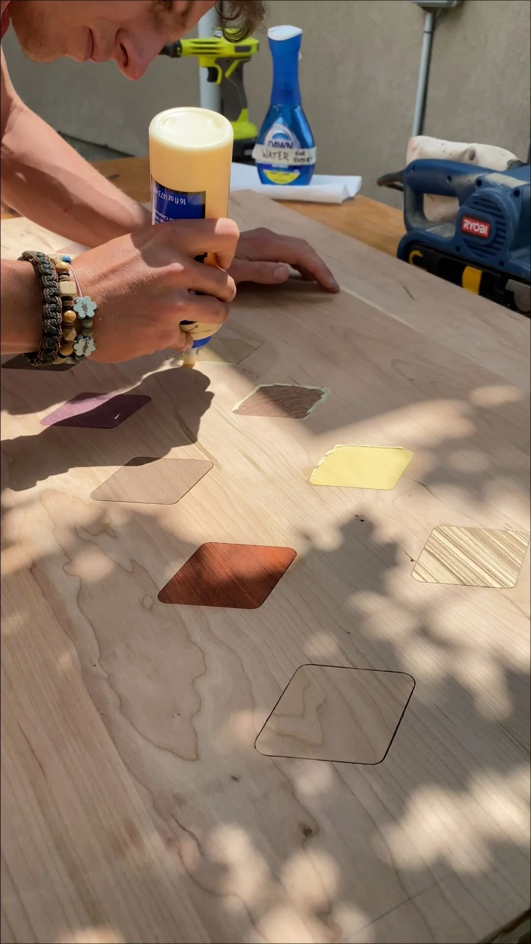 A person is using a glue gun to glue colored acrylic or wooden pieces onto a wooden surface. The surface has various cut-out shapes filled with different colors and textures, creating a geometric design.