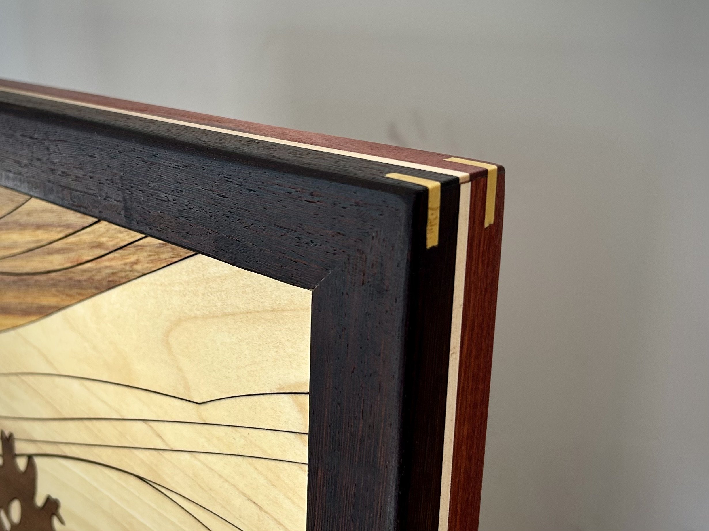 Close-up of a corner of a wooden table or furniture piece with multicolored wood inlay details, set against a plain gray wall.
