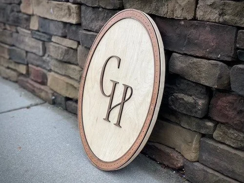 A round wooden sign with the letters 'CH' engraved, leaning against a stone wall.
