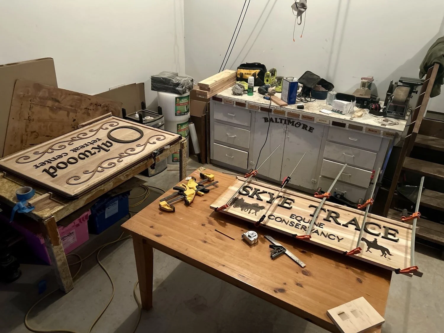 Workshop with wooden signs being assembled and painted, including one with the text 'Skyfire Equine Convalescence' and another with 'Arlington' and decorative elements, clamps, tools, and supplies.