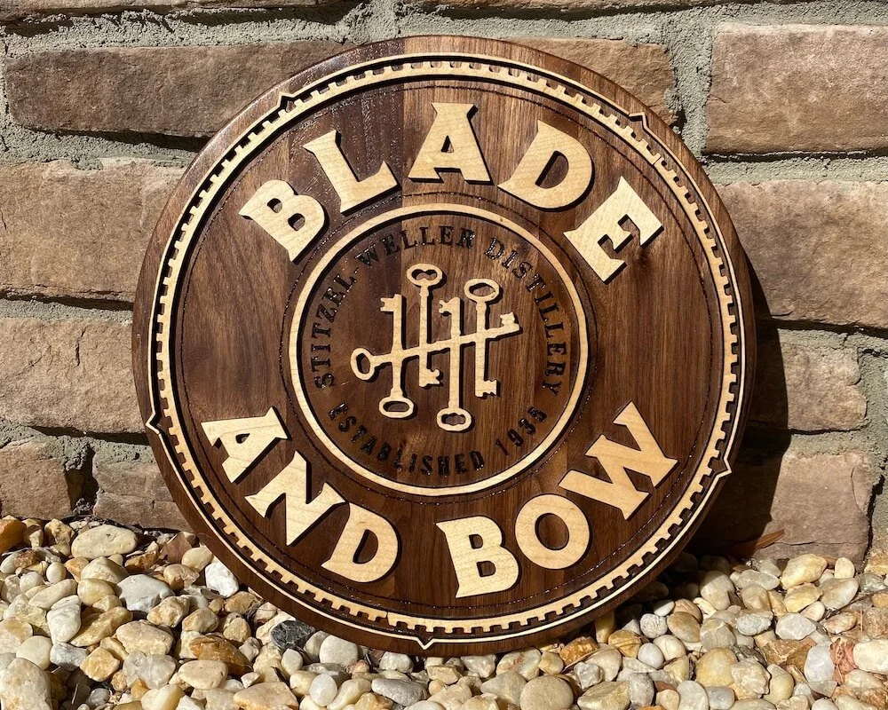 A round wooden sign with the words "BLADE AND BOW" and the phrase "STILTZER WELDER DISTRIBUTED SINCE 1936" around a central logo of interconnected keys, leaning against a brick wall and resting on a bed of small rocks.