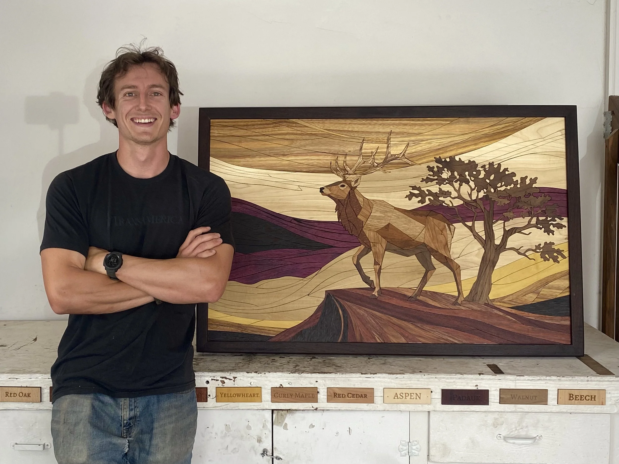 A young man with curly brown hair, wearing a black T-shirt and a wristwatch, standing in front of a large wooden art piece depicting a deer, trees, and a landscape with colorful layers.