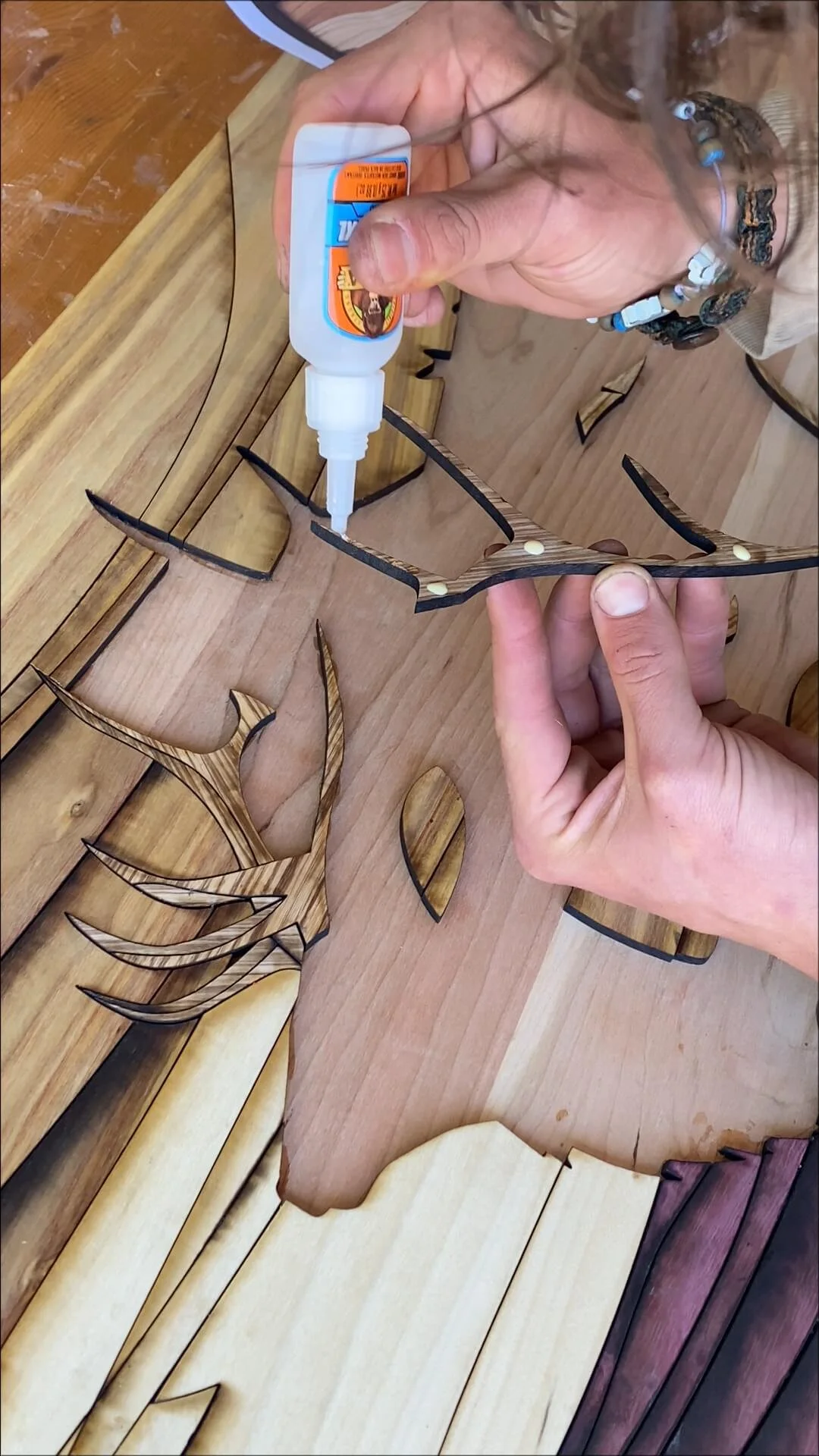 Close-up of a person applying glue to the back of a wooden cutout shaped like a wolf or fox head. Various other wooden shapes and cutouts are on a wooden surface.