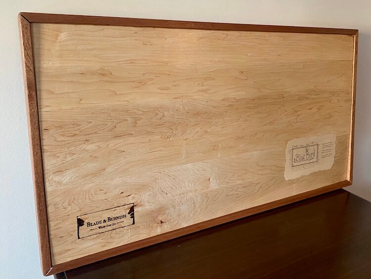 The back of a maple wood sign with mahogany framing, showing the Blade & Burnish logo burnt in and laminated design blueprints.