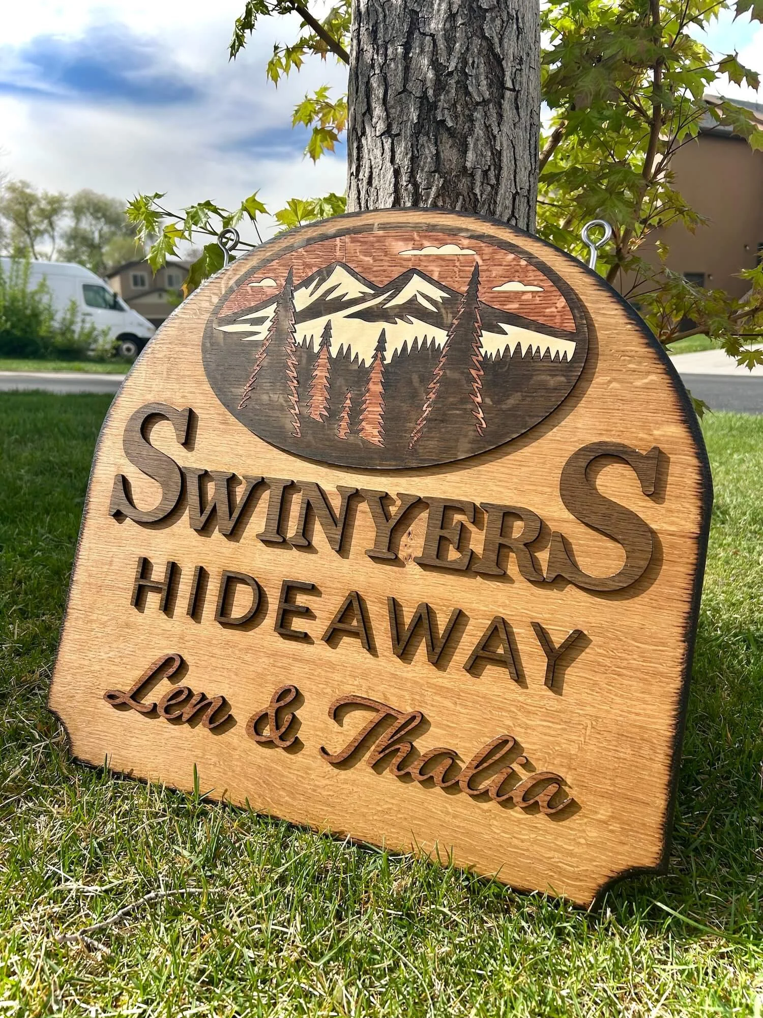 A wooden sign with a mountain and forest design, partially visible text 'SWINYER'S HIDEAWAY, Len & Thackera,' leaning against a tree in a grassy area, with houses and a van in the background under a cloudy sky.
