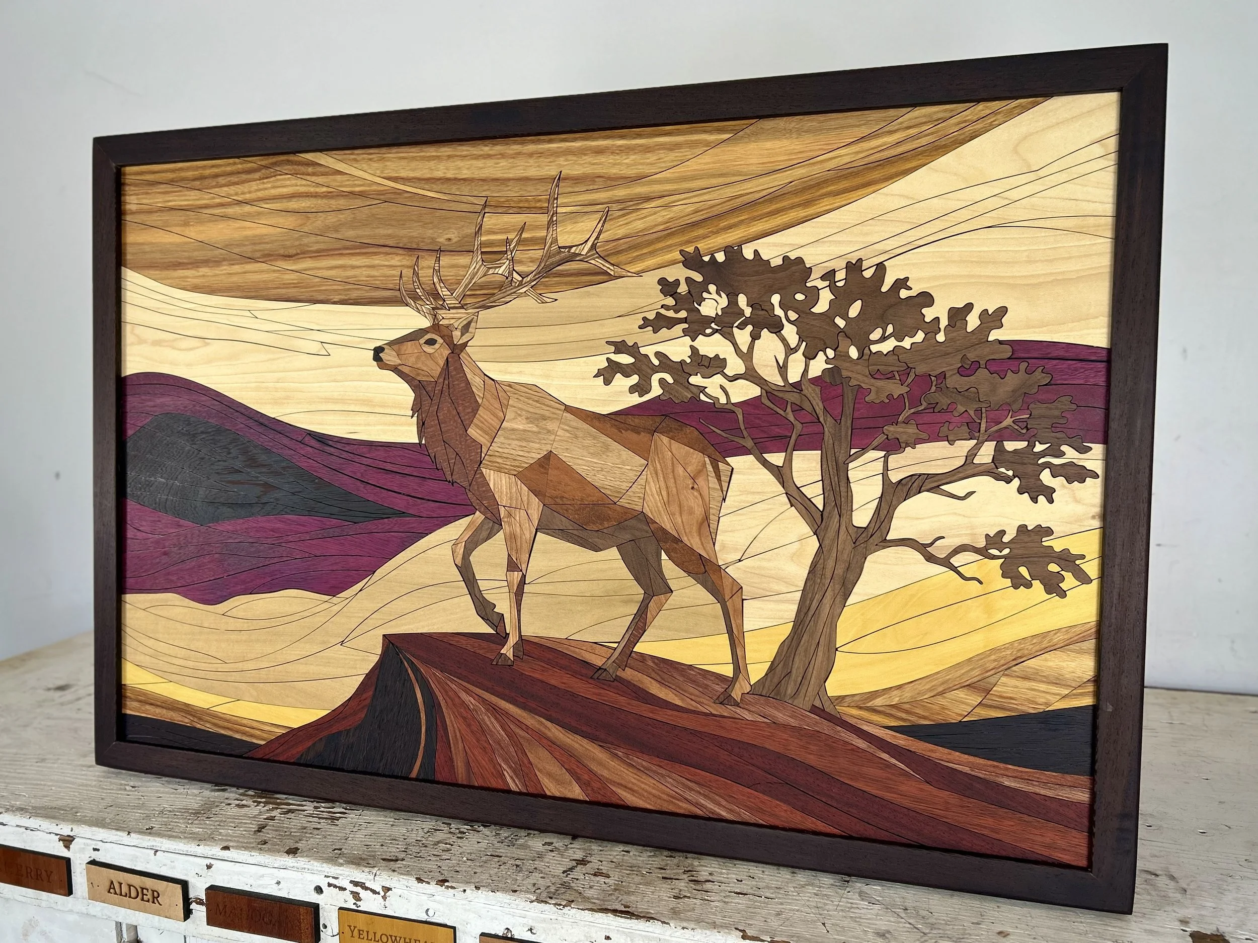 Wooden framed artwork of a deer standing beside a tree, with mountainous landscape and sky in the background, created using different shades of wood.