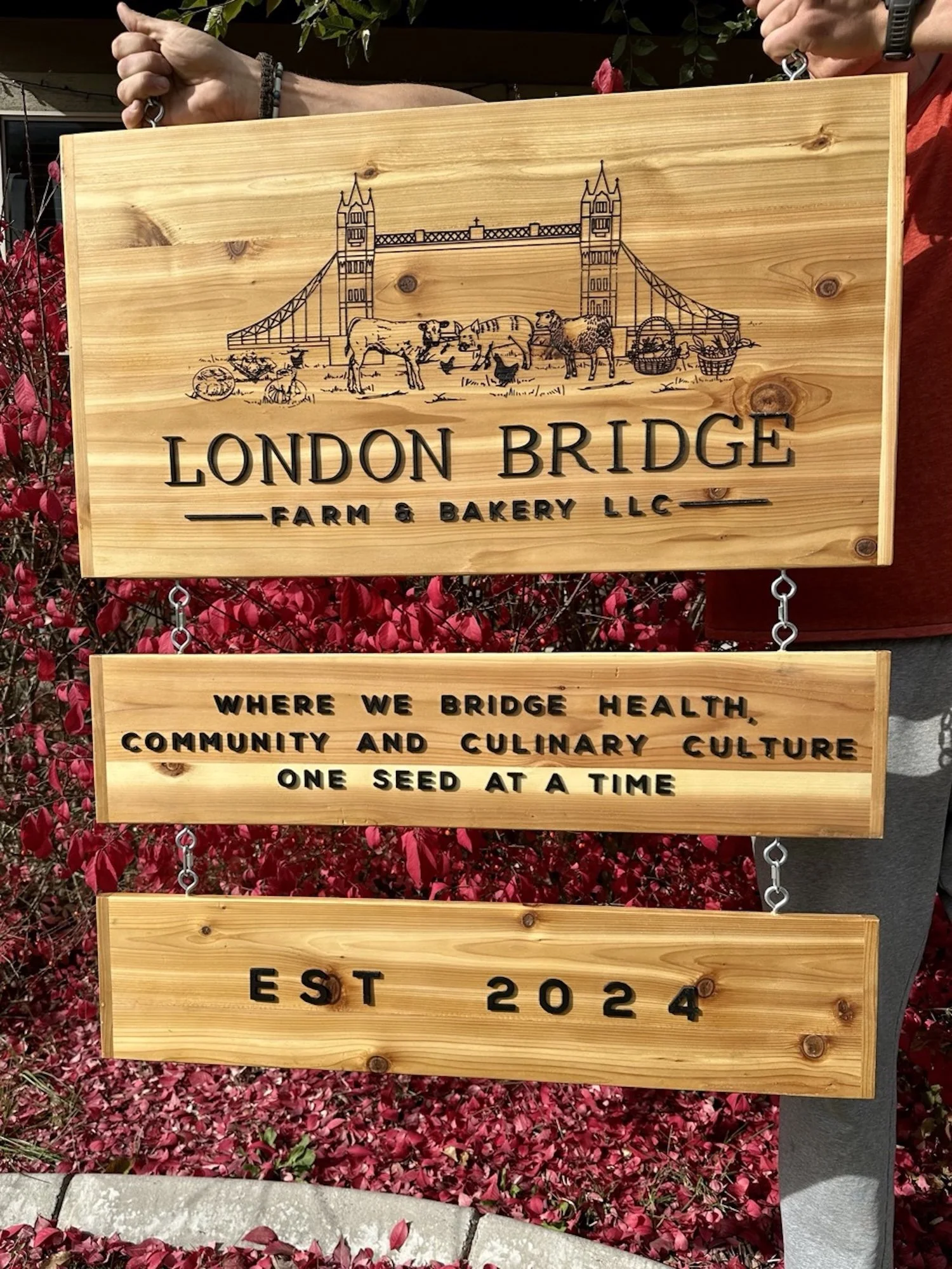 Wooden sign for London Bridge Farm & Bakery LLC featuring an illustration of London Bridge with farm animals including cows, chickens, ducks, and a rabbit, and decorative baskets under the bridge.