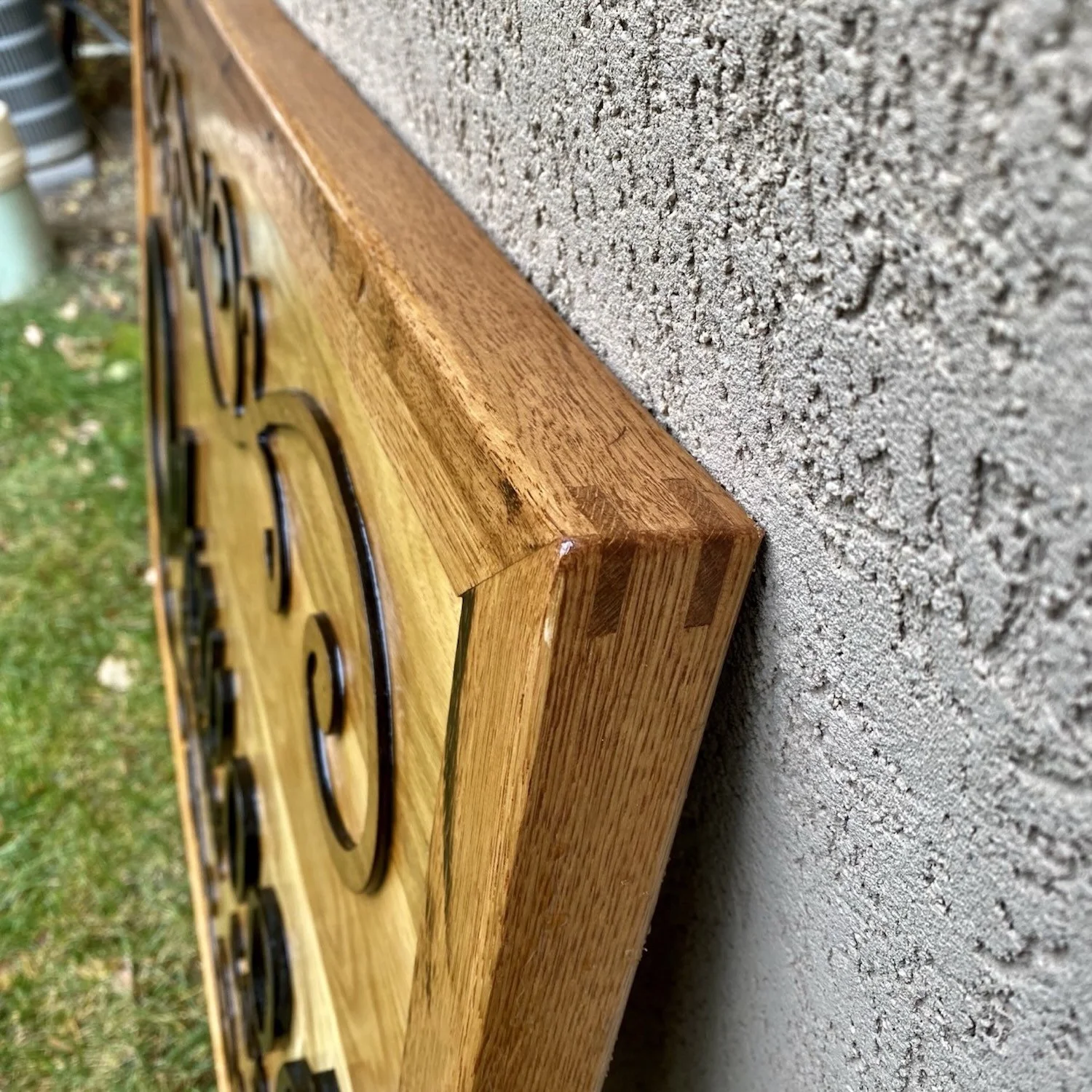 Close-up of a wooden sign with raised black lettering, mounted on a textured concrete wall, next to a grassy area with leaves.