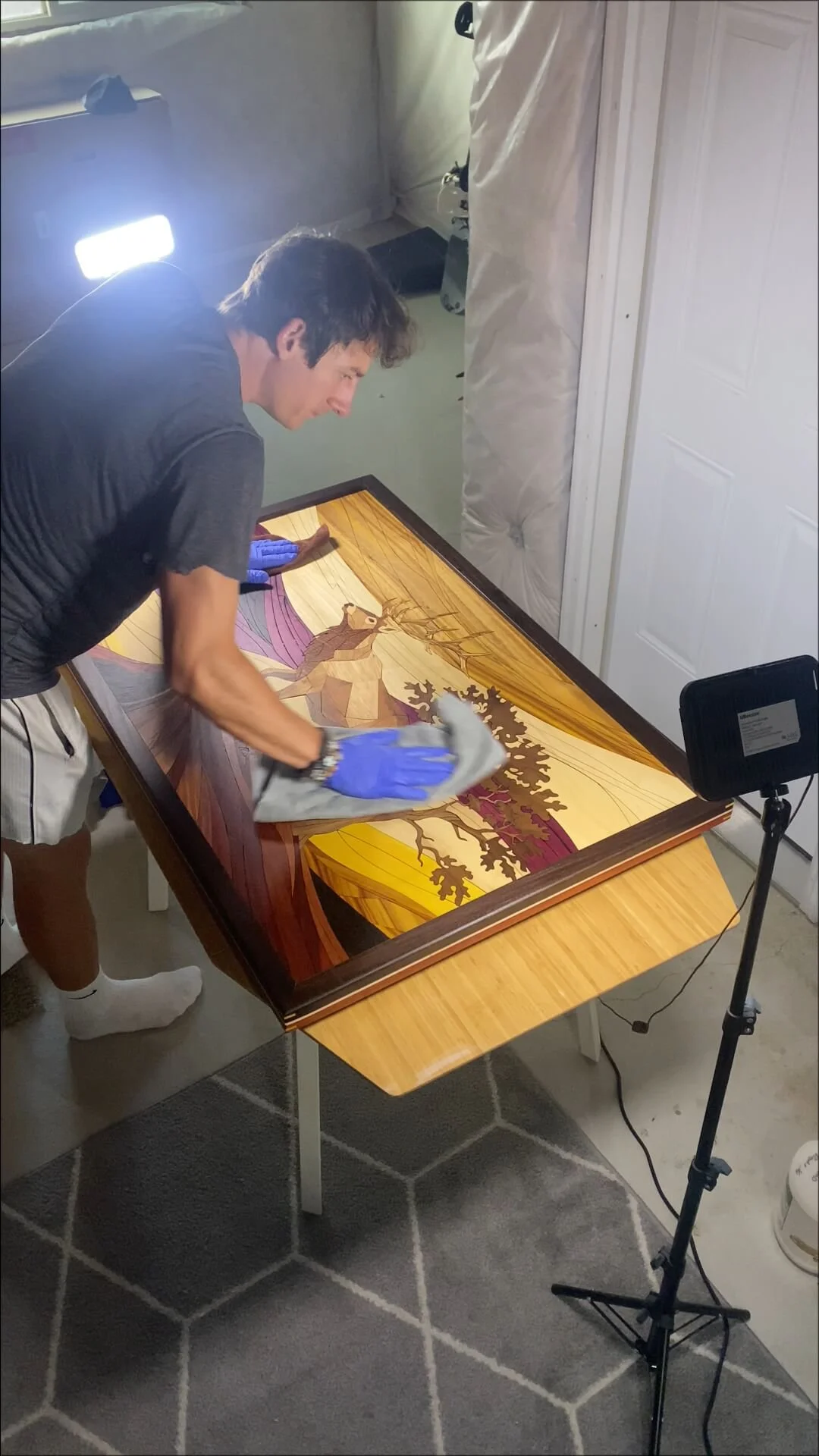 A person is cleaning a framed wooden landscape art piece on a table, wearing gloves, with a bright light and a camera or light stand nearby.