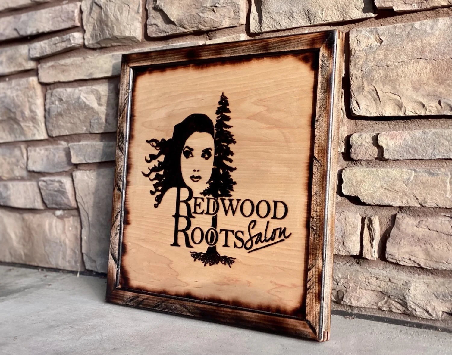 Wooden sign with a black silhouette of a woman's face, pine tree, and the text 'Redwood Roots Salon' in black, leaning against a brick wall.