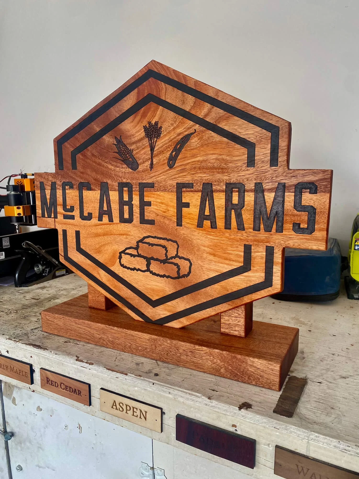 Wooden sign with black lettering that reads 'McCabe Farms' and has illustrations of a corn cob, wheat, and a pod or chili pepper.