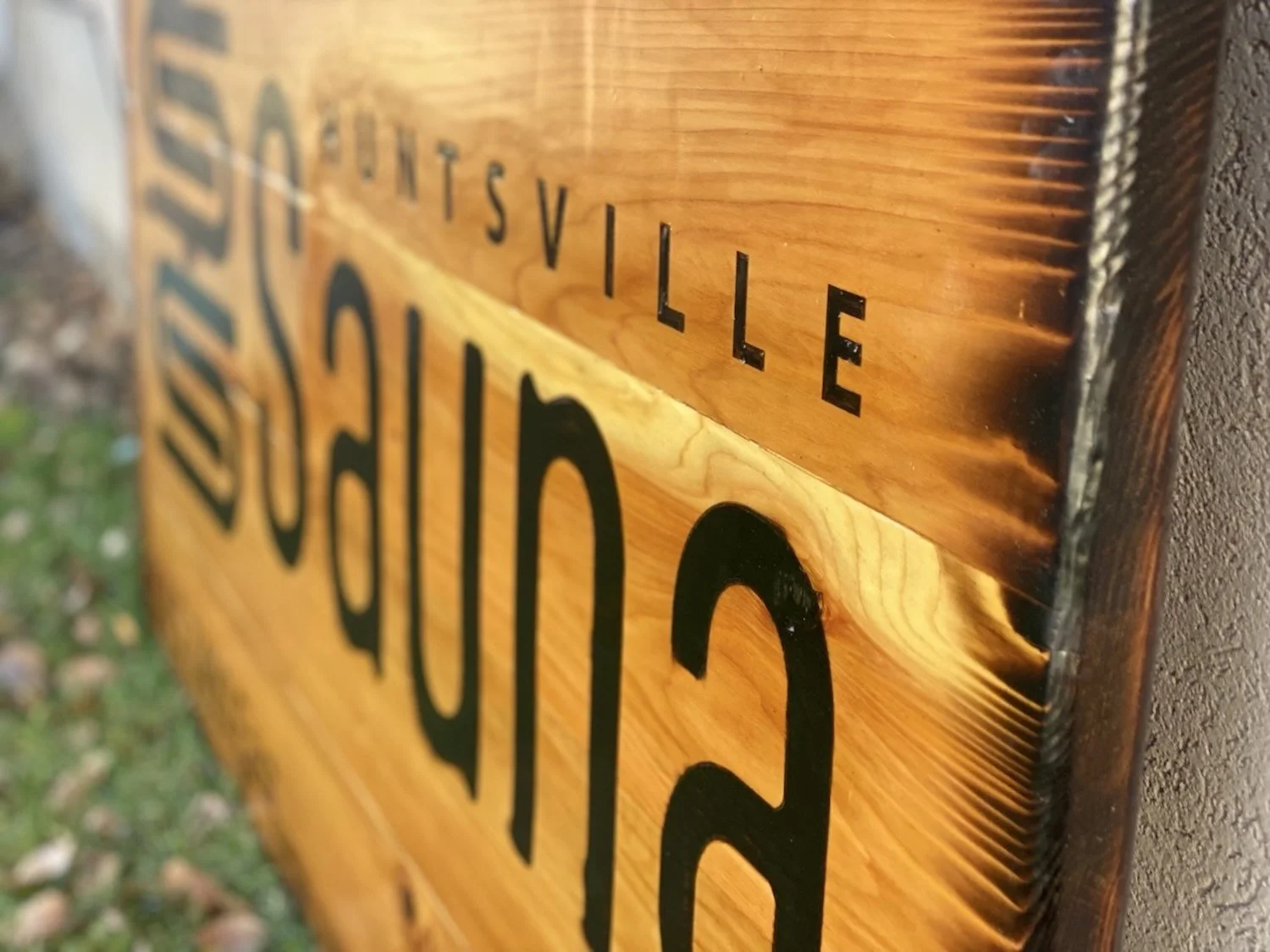 Close-up of a wooden street sign with black burnt-in text, showing part of the word '"SAUNA" and other words above it, mounted on a textured wall.