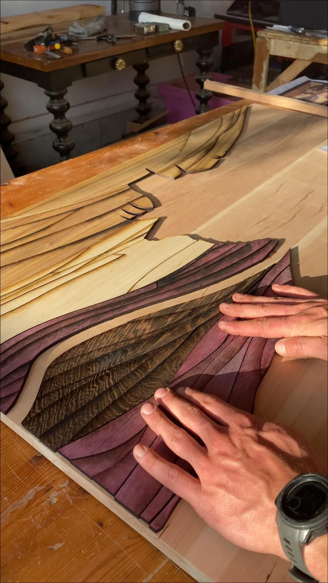 Hands arranging multicolored wood veneer sheets on a wooden table, with a workshop background visible.