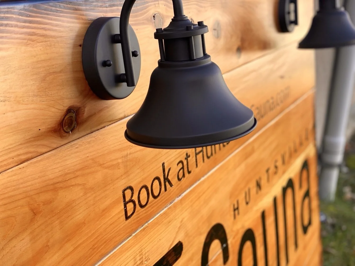 Close-up of black outdoor light fixture mounted on wooden cedar sign with partial visible sign reading 'Book at H...'