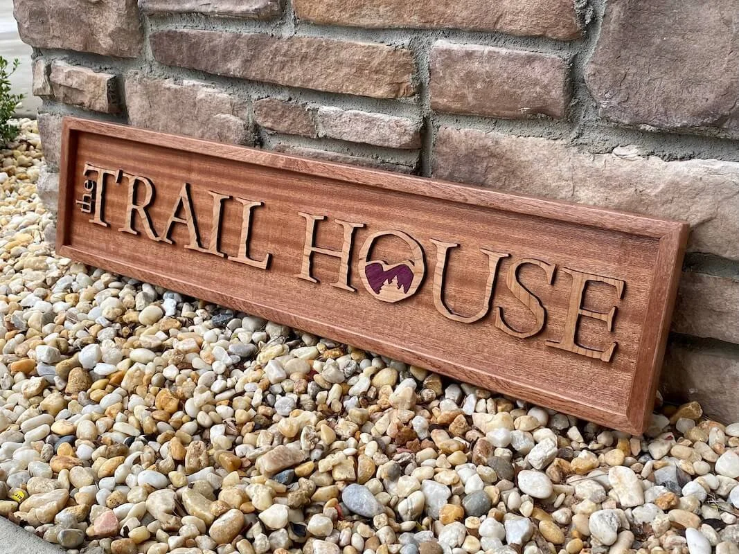 Wooden sign with the words 'TRAIL HOUSE' and mountain and sun logo, leaning against a brick wall, on a bed of small multicolored rocks.