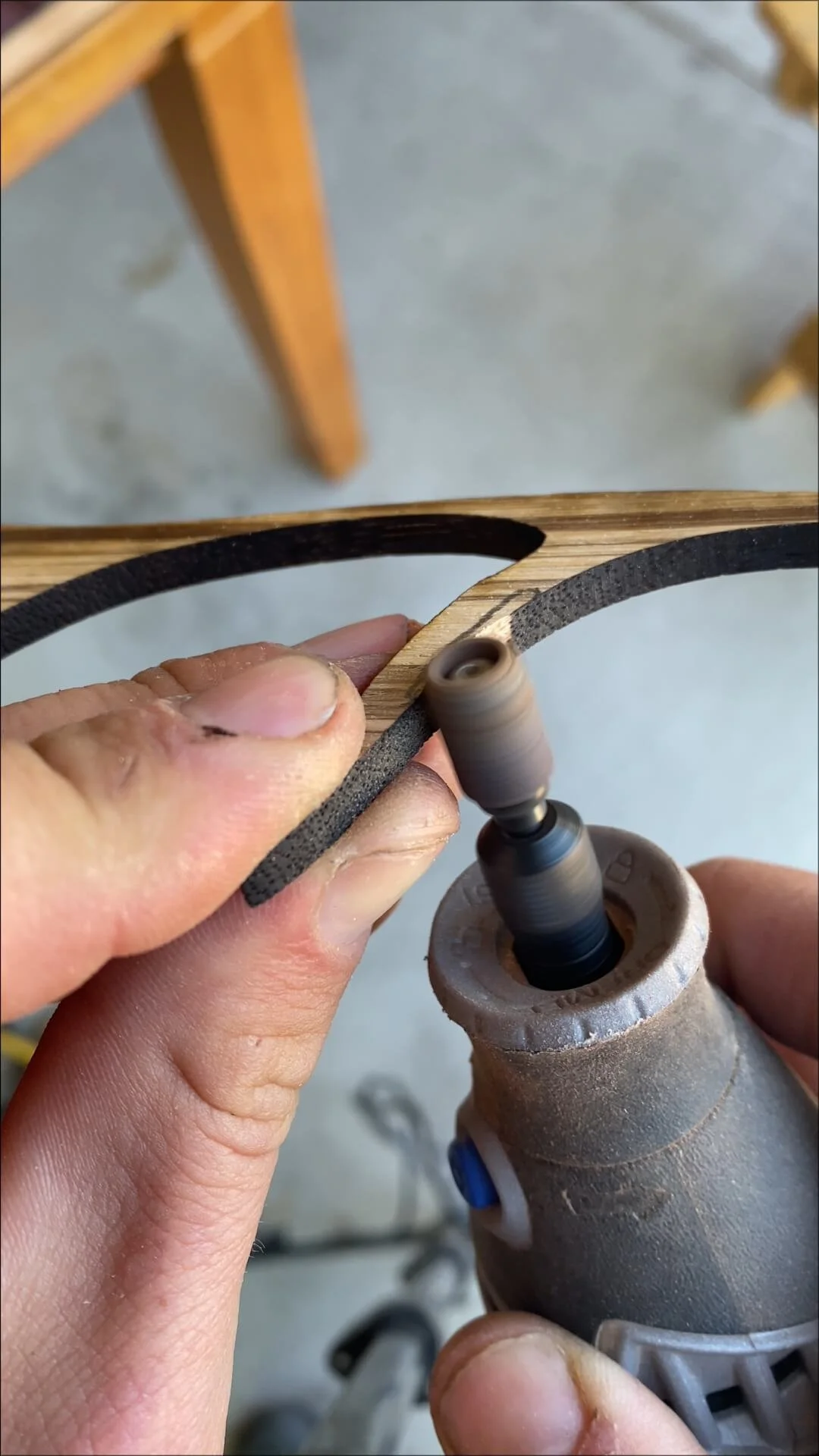 Close-up of a person using a rotary tool to sand a thin strip of dark-colored wood or material, with a wooden piece in the background.