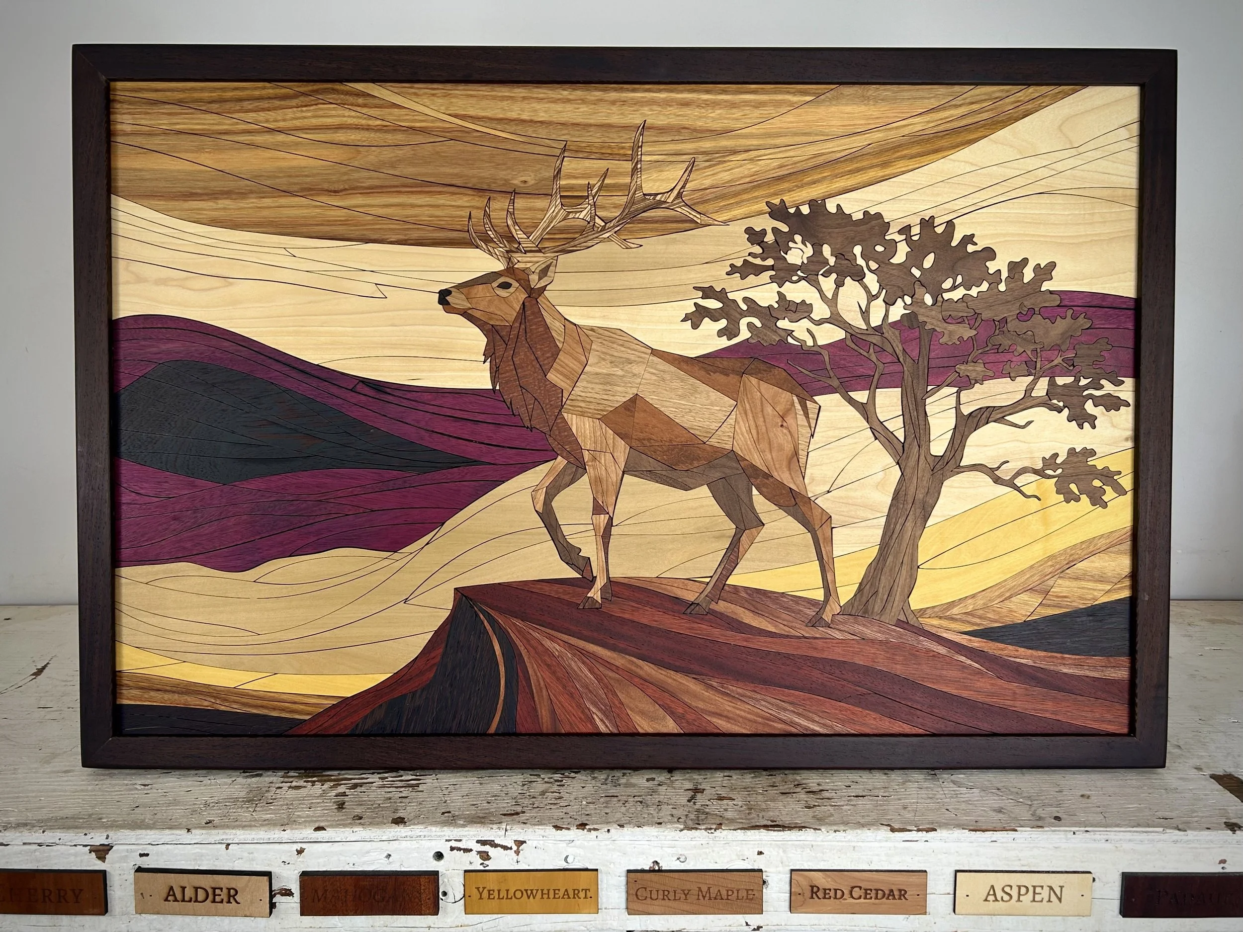 A wooden art piece depicts a deer standing on a rocky outcrop, with a tree to its right and colorful hills in the background, framed in a dark border.