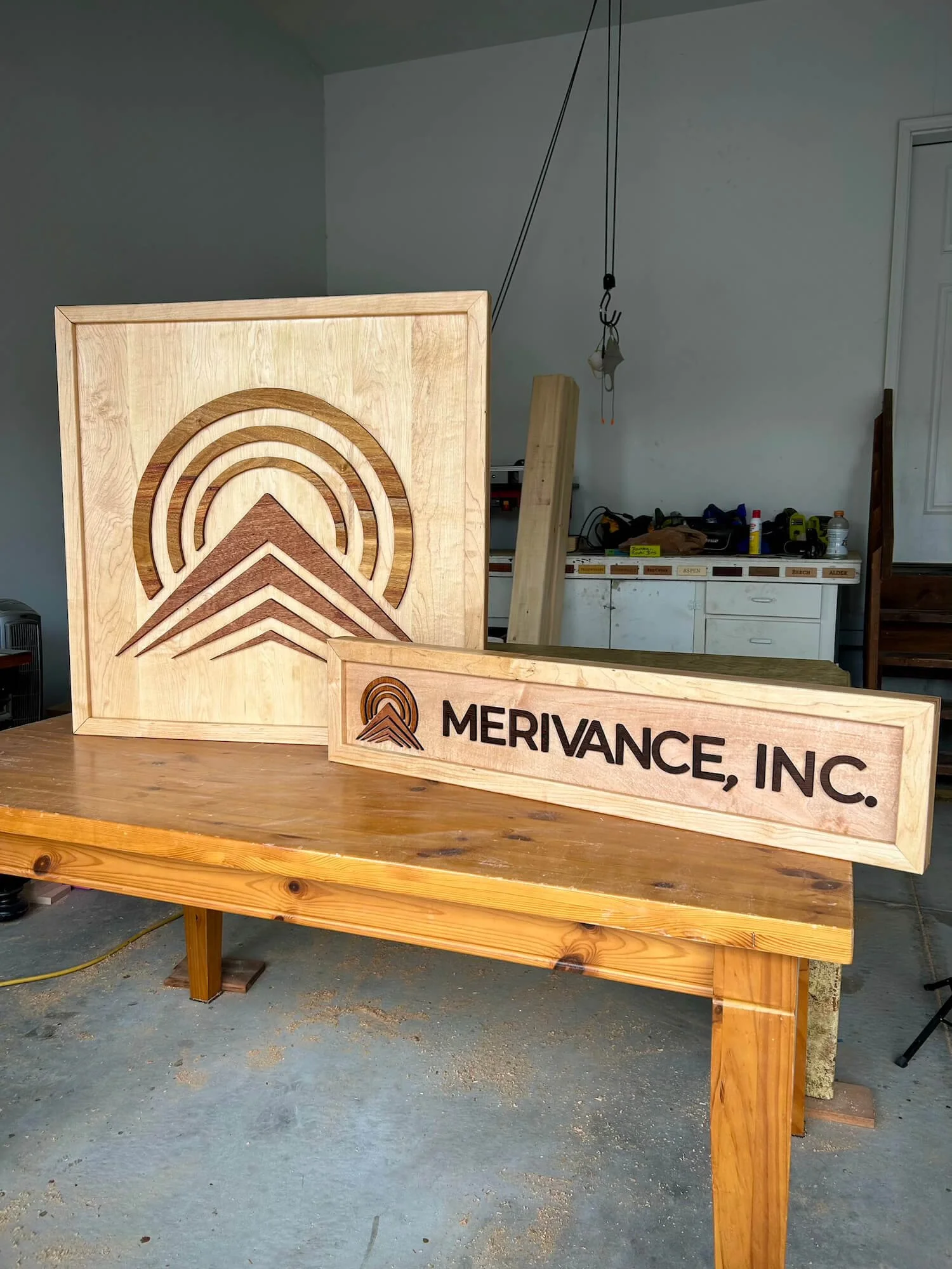 Wooden sign with the logo and name 'MERIVANCE, INC.' on a workshop table, with a large wooden panel featuring a carved logo in the background.