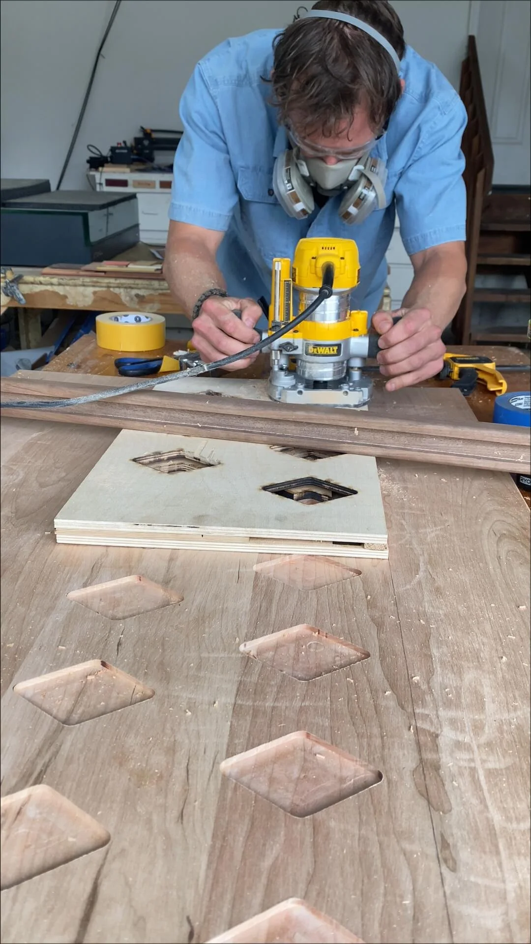A man wearing safety goggles, a mask, and headphones is using a woodworking router on a piece of plywood with diamond-shaped cutouts. Several cutouts are visible on the table and in the plywood.