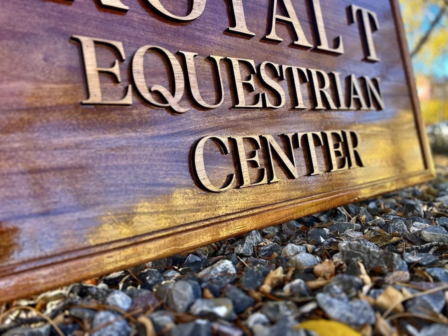Close-up of a wooden sign with carved letters reading 'Equestrian Center,' placed on rocks with blurred trees and sky in the background.