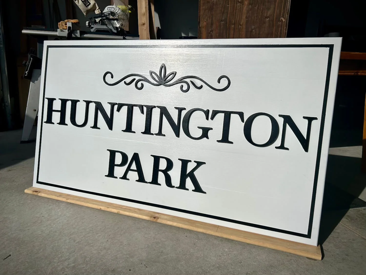A white sign with black border and text reading "Huntington Park" on a paved surface, with items like a helmet, saw, and tools on a workbench in the background.