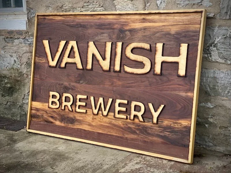 Wooden sign reading 'VANISH BREWERY' with carved letters, leaning against a stone wall.
