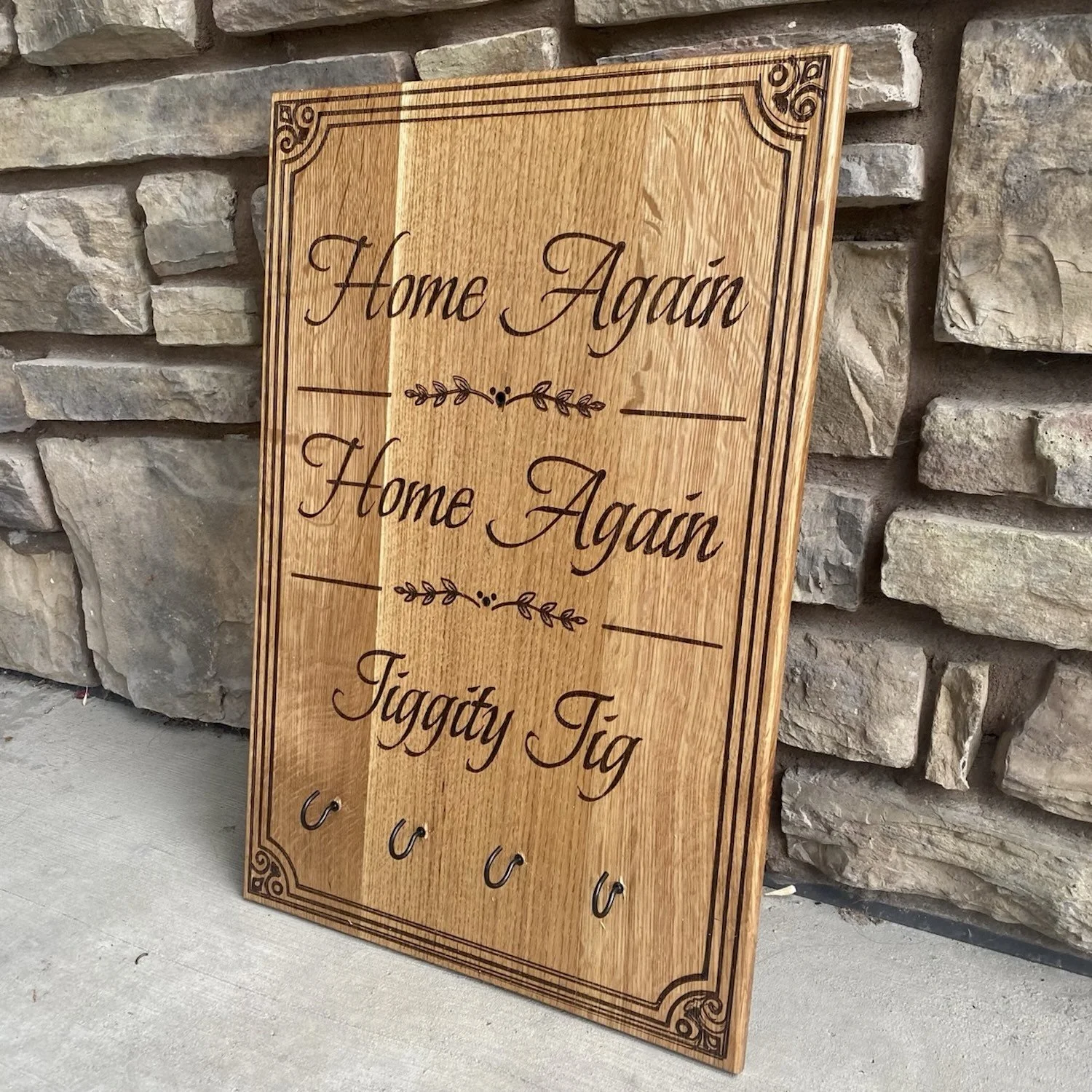 Wooden decorative sign with a quote about home, featuring a list of names or words, accompanied by decorative lines and corner designs, leaning against a stone wall.