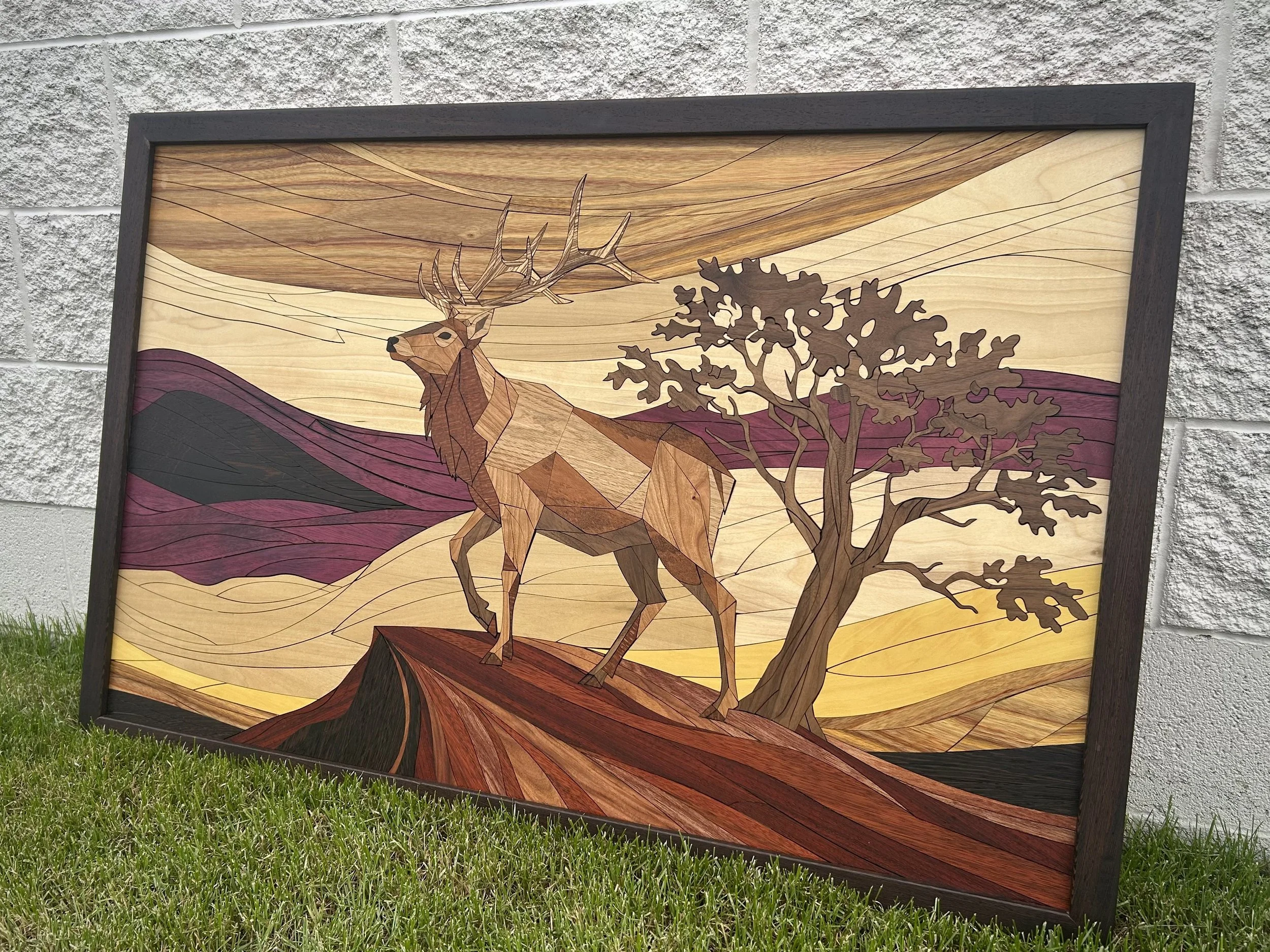 A framed artwork of a deer standing by a tree, created with layered wood in a landscape scene, mounted on a brick wall outdoors.
