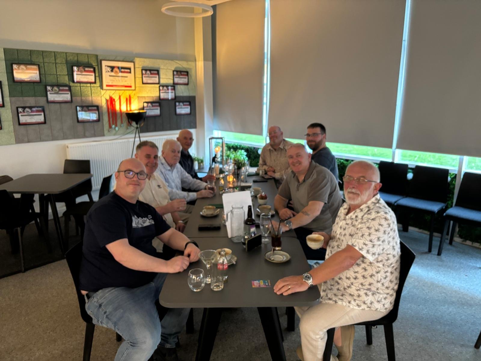 A group of nine men sitting around a rectangular table in a restaurant, smiling for the photo. The table has drinks and small plates on it. The background features a wall with framed pictures and a window with closed shades.