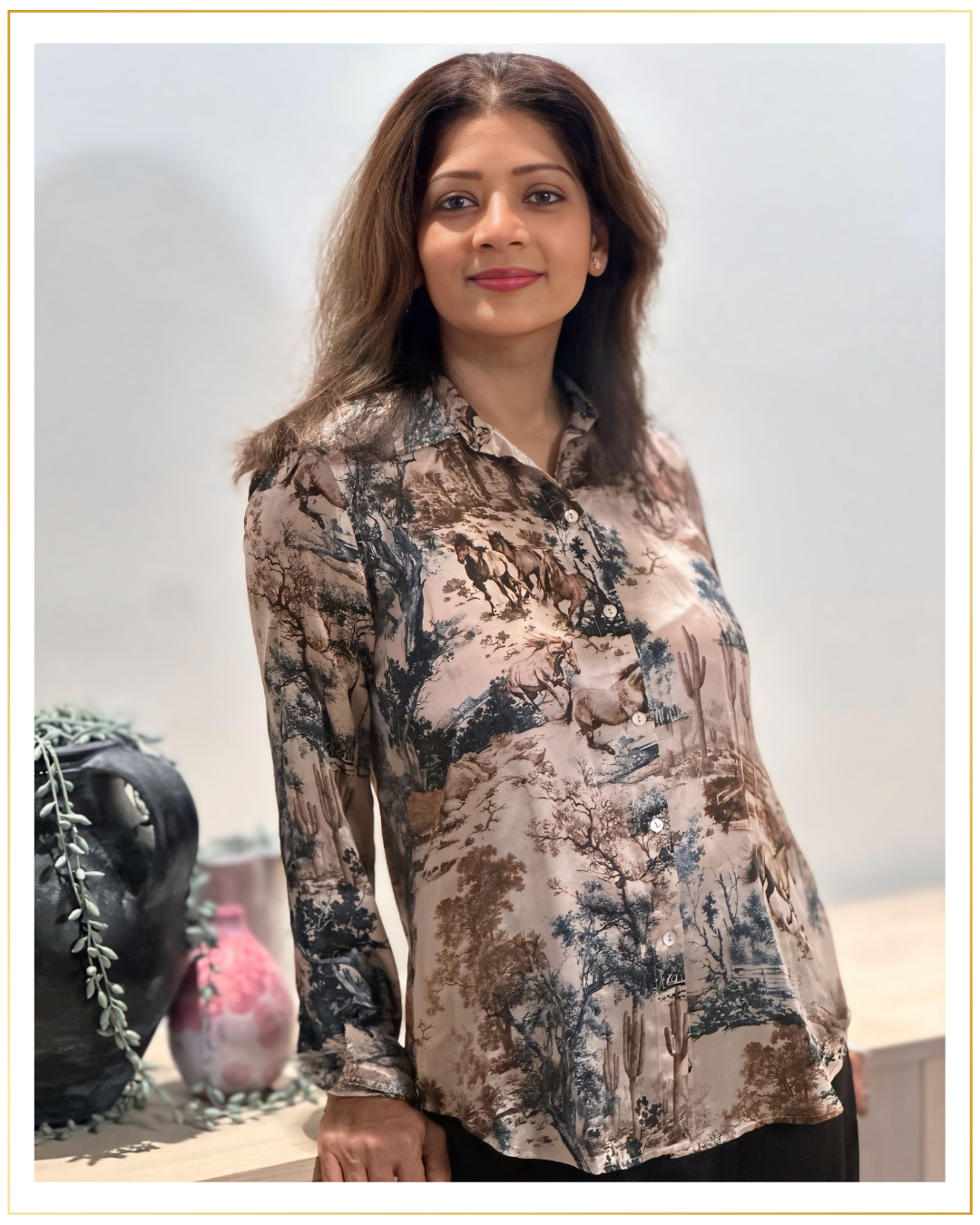 A woman with shoulder-length brown hair, wearing a patterned blouse with a nature scene featuring trees, animals, and desert landscape. She is standing in front of a plain white background with a decorative gold border, next to vases and plants on a wooden surface.