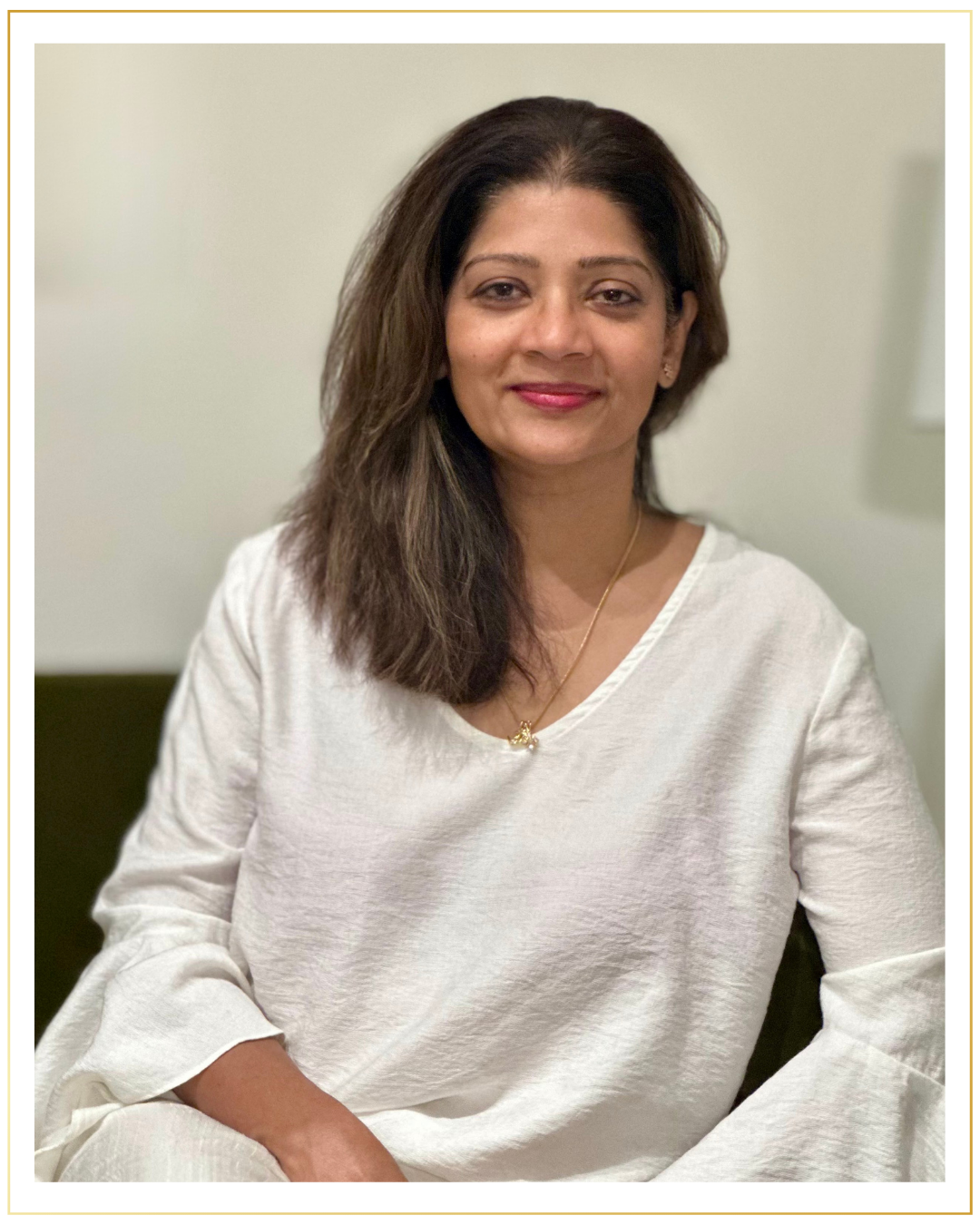 A smiling woman with shoulder-length brown hair, wearing a white top and a gold necklace, sitting on a green chair against a neutral background.