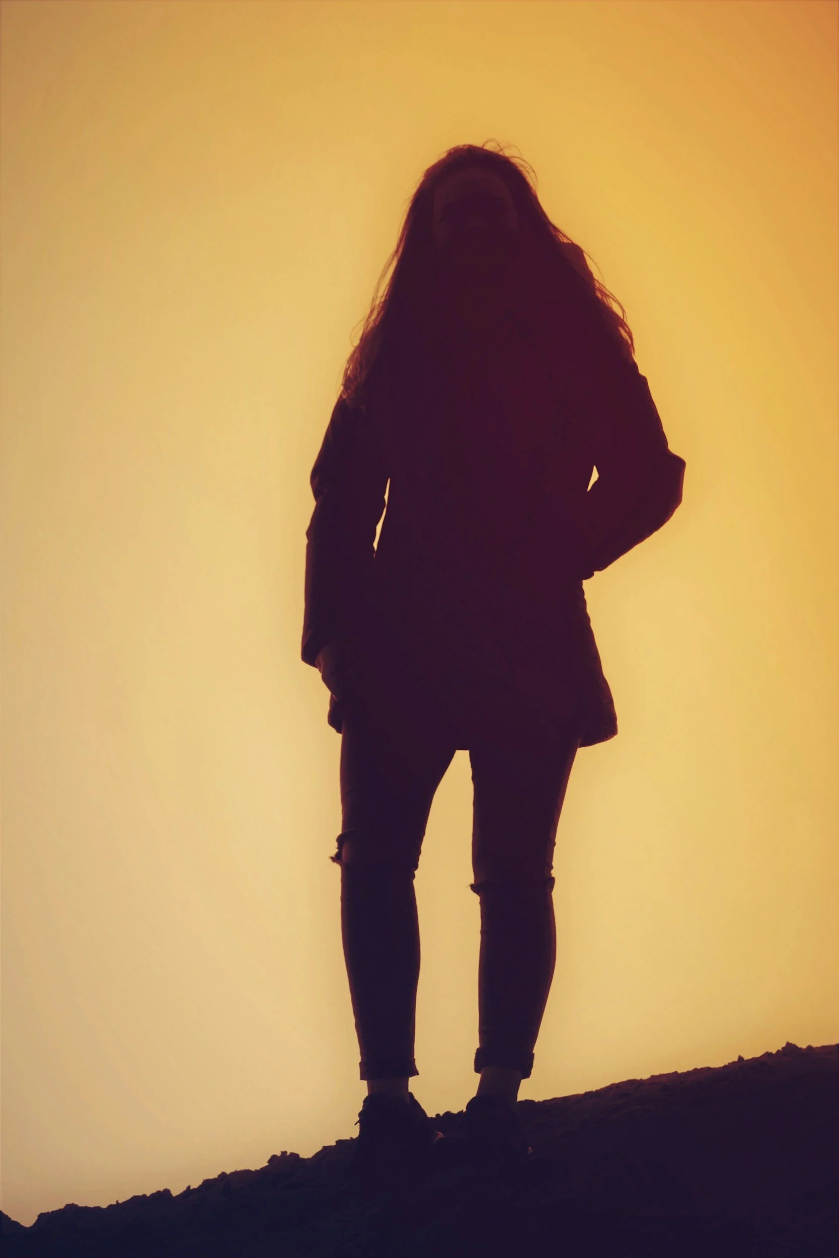 Silhouette of a woman standing on a hill at sunset or sunrise with a yellow-orange sky in the background.